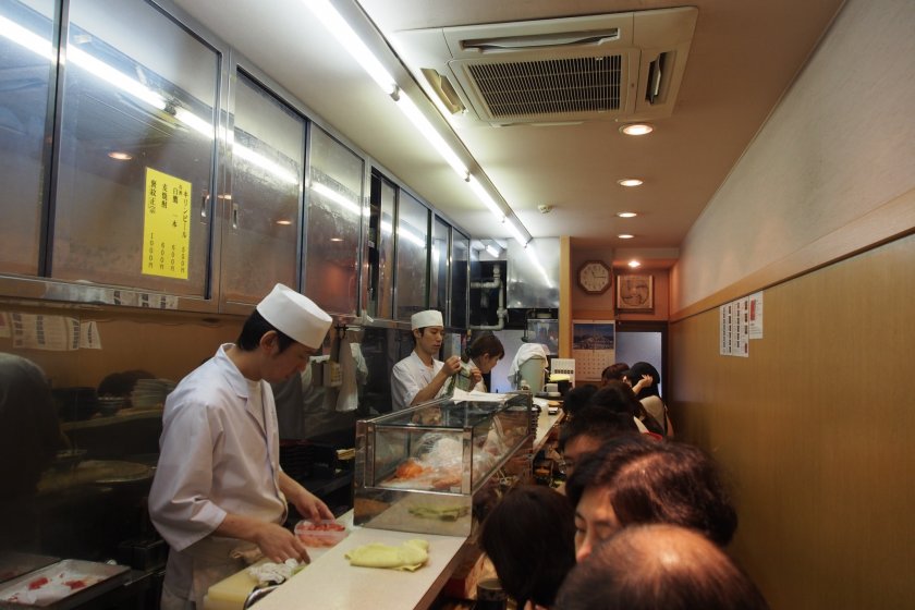 Bên trong một trong những nhà hàng ở Chợ Cá Tsukiji; mọi người ngồi ngay ngắn trong khi các đầu bếp chuẩn bị bữa ăn