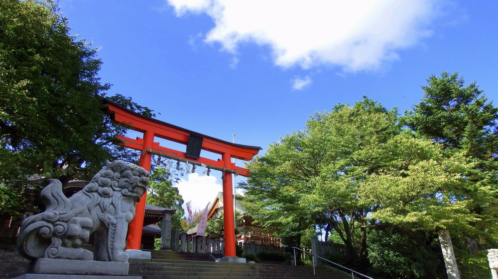 Entrance of Fujishima Shrine on the hillside of Mt. Asuwa. The red torii gate and green trees under the blue sky make a striking contrast.