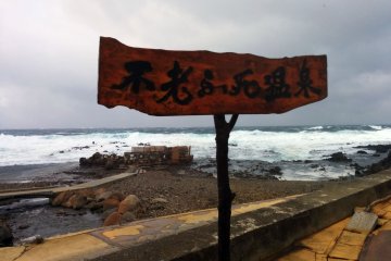 A sign reads Furofushi Onsen. That ramshackle shelter in the background that looks like it will be washed away by the next big wave is your destination.